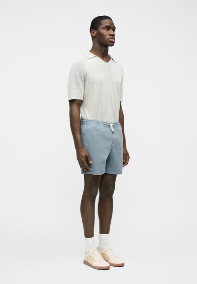 Jeune homme debout, portant un pull à manches courtes beige clair, un short bleu à cordon, des chaussettes blanches et des baskets beiges, sur fond uni.