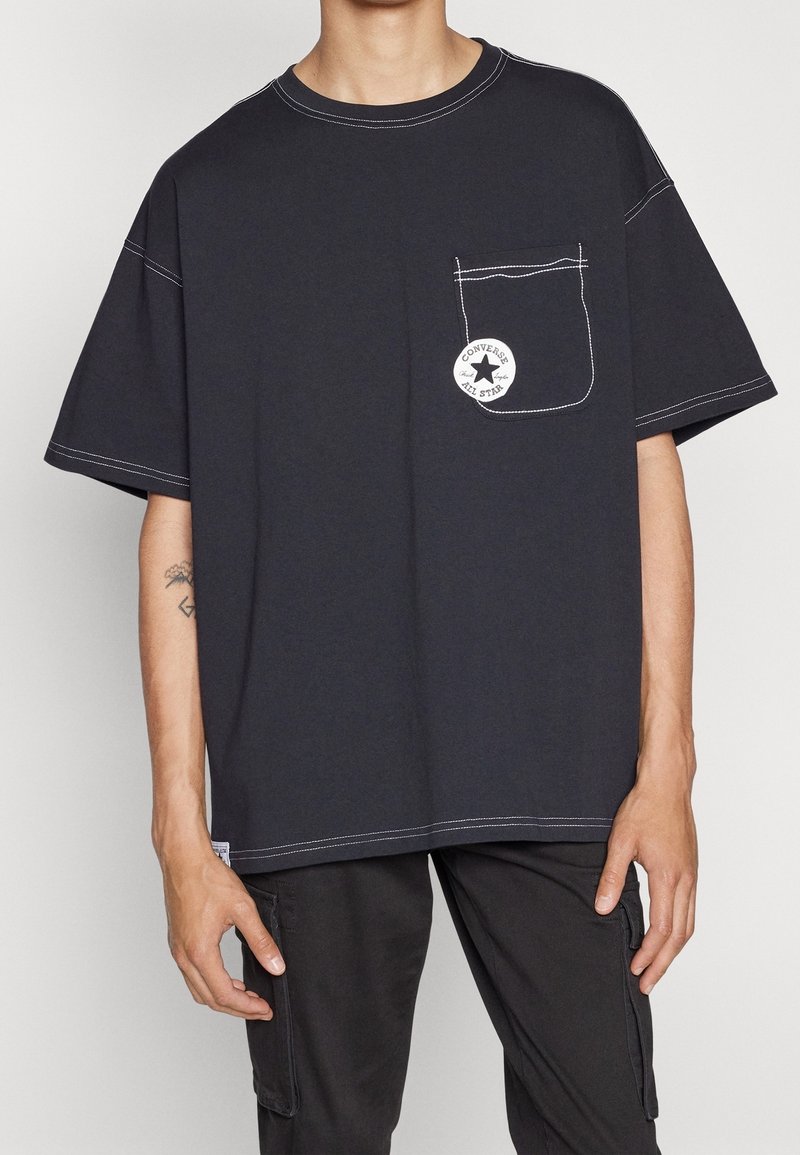 Personne portant un T-shirt noir oversize avec des coutures blanches et une poche poitrine affichant un écusson rond du logo Converse All Star.