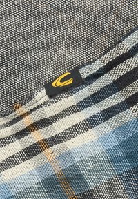 Tissu tissé avec un motif à carreaux gris et bleu, avec une étiquette noire portant un logo jaune. Surface texturée avec des nuances de couleur variées.