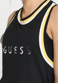 Svart mesh linne med gula och vita randiga detaljer, med en reflekterande silverlogotyp "GUESS" över bröstet.