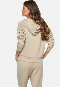 Beige hoodie met een trekkoordcapuchon, ribbels aan de mouwen en onderkant. Zacht materiaal en een relaxed fit, gecombineerd met bijpassende broek. Achteraanzicht getoond.