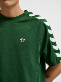 Personne portant une chemise verte à manches courtes avec des bandes blanches en chevron sur les épaules et un petit logo brodé sur la poitrine.