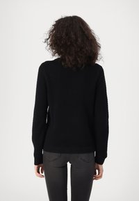 Svart strikket genser med et strukturert mønster, ribbestrikkede mansjetter og hem, vist bakfra, kombinert med mørkegrå jeans.