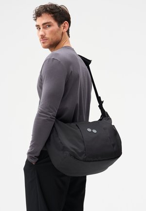 Mann trägt graues Langarmshirt und schwarze Hose und hat eine große schwarze Umhängetasche mit zwei runden Knöpfen über der Schulter.