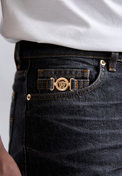 Jeansi din denim închis cu elemente de hardware metalic auriu, un emblema proeminentă cu leu pe talie și detalii de cusătură portocalie contrastantă.