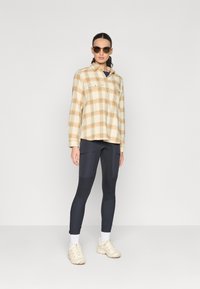 Camicia di flanella a quadri beige e arancione con tasche frontali, abbinata a leggins neri e calzini alla caviglia bianchi, completata da sneakers beige e occhiali da sole.