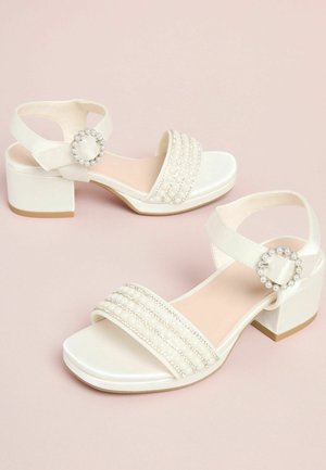 Valged madala kontsaga plokk-sandaalid pärlite ja kivikestega kaunistatud esirihmaga ning pärliklambri ja pahkluurihmaga, heleroosa tausta peal.
