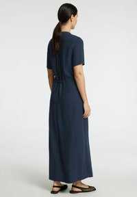 Robe t-shirt longue bleu marine avec des manches courtes, ceinture élastique et silhouette fluide. Portée avec des chaussures plates ouvertes noires.