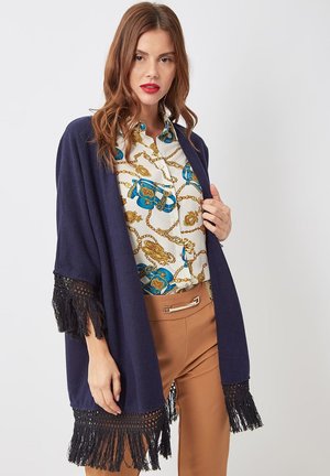 Mujer con un chal de flecos azul marino, blusa blanca con estampado de cadenas doradas y azules, y pantalones beige con cinturilla acentuada en metal.
