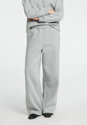 Person trägt hellgraue Sweatpants mit weitem Bein und elastischem Bund sowie schwarze Schuhe, Arme verschränkt, vor weißem Hintergrund.