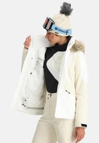 Giacca da sci bianca con accentuazioni color crema chiaro, cappuccio foderato in pelliccia, tasche con zip e etichette interne. Indossata sopra uno strato base nero.