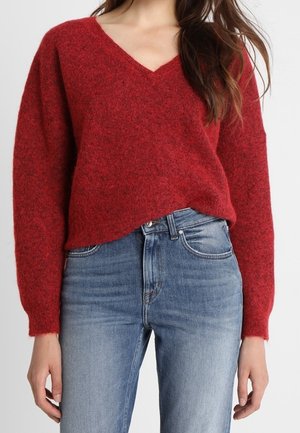 Kvinna som bär en röd V-ringad fluffig tröja instoppad i ljusblå högmidjade jeans mot en enkel bakgrund.