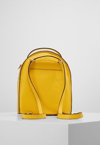 Mochila de cuero amarillo con textura suave, diseño curvado, ribetes negros en contraste y correas ajustables. Vista trasera, con hardware mínimo visible.