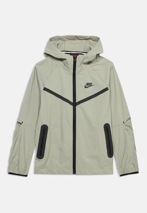 Jasnobeżowa kurtka z kapturem Nike z czarnym zamkiem błyskawicznym, czarnymi bocznymi kieszeniami na zamek oraz czarnym wzorem w kształcie szewronu na klatce piersiowej i rękawach.