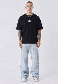 Black cotton t-shirt with central white logo, paired with light blue wide-leg jeans and white sneakers, featuring a relaxed fit and casual design.