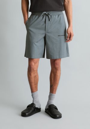 Man die middellange grijze shorts met trekkoord, grijze sokken en zwarte instapschoenen draagt, staand tegen een effen achtergrond.
