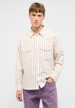 Junger Mann, der ein cremefarbenes Hemd mit vertikalen braunen Streifen und lila Hose trägt, steht mit einer Hand in der Tasche vor einem einfarbigen Hintergrund.