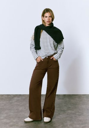 Chemise à rayures à manches longues grise et blanche, pantalons larges marron foncé avec des accents de boutons métalliques, écharpe en tricot noire drapée sur les épaules.