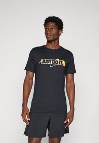 Sort bomulds T-shirt med "JUST DO IT" grafik i gul og hvid, sammen med et Nike-logo, parret med sorte sportshorts.