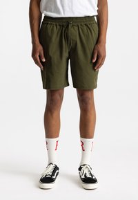 Olijfgroene shorts met een elastische tailleband en trekkoord. Gepaard met zwarte sneakers en witte sokken met rode letters.