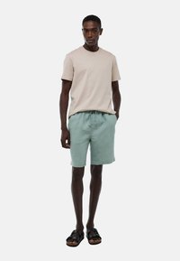 T-shirt en coton beige associé à un short vert clair, doté d'une taille élastique. Des sandales noires complètent le look. Design simple et décontracté.