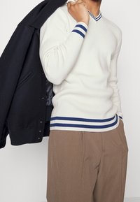 Hvid strikket sweater med marineblå stribet ribkrave og manchetter, parret med brune plisserede bukser. En sort jakke er slængt over den ene skulder.