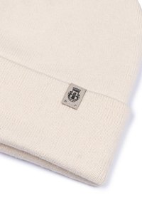 Light beige knit beanie, soft texture, ribbed design, featuring a small gray tag with an emblem. Simple and functional.
