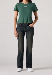 Jean évasé bleu foncé avec un léger effet délavé, associé à un t-shirt court vert avec un logo blanc Levi's sur la poitrine. Baskets blanches.