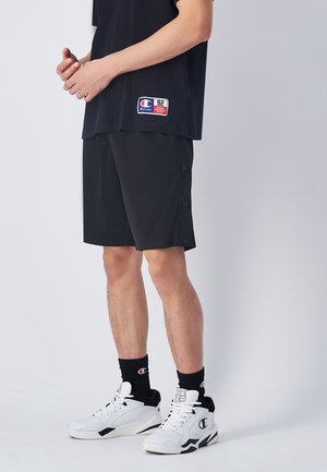 Sort shorts med sidelukningsdetaljer, parret med en sort t-shirt med et logo-patch. Båret med hvide og sorte sneakers.