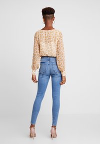 Halenka s dlouhým rukávem a květinovým vzorem v světle žluté a bílé barvě, zkombinovaná s džínami obepínajícími postavu v modré barvě a nahými podpatky; hladká textura, ležérní styl.