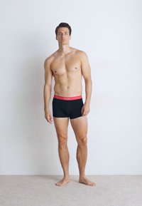Fekete boxer alsónadrág piros derékkal, testhezálló kialakítással. A modell semleges pózban áll egy egyszínű háttér előtt.