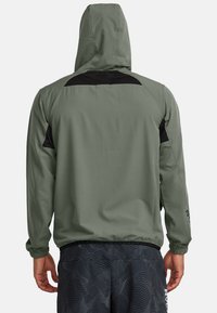 Chaqueta ligera con capucha en verde oliva, que cuenta con acentos de malla negra en los hombros, puños elásticos y un cordón en el dobladillo.