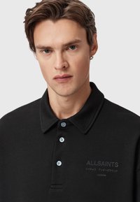 Ung mand med kort brunt hår iført en sort AllSaints polo-shirt med knapper og et lille broderet logo på brystet.
