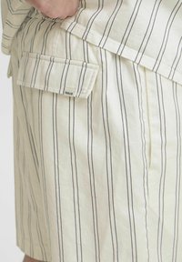 Hvite stoffshorts med smale blå vertikale striper, med en sidelomme og etikett merket 'SOLID'. Teksturen ser myk og uformell ut.
