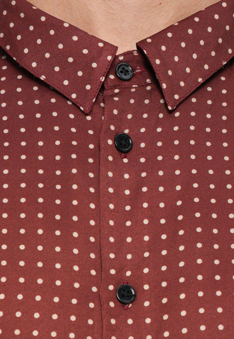 Bordeaux knoopshirt met een puntkraag, voorzien van een klein wit stippenpatroon en zwarte knopen. Zachte stof met een gladde textuur.