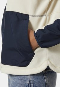 Mikina z fleece materiálu s krémovým telom, námorníckymi rukávmi a elastickými manžetami. Má bočnú kapsu a prešívané detaily, kontrastné textúry.