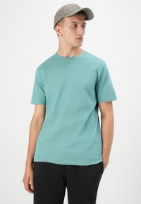 Τιρκουάζ βαμβακερό t-shirt με κοντό μανίκι και στρογγυλή λαιμόκοψη, συνδυασμένο με σκούρες παντελόνες, φορώντας από ένα μοντέλο που κοιτάει ελαφρώς αριστερά.