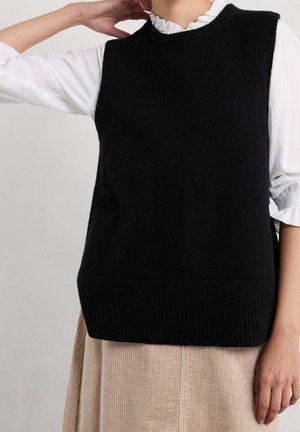 Personne portant un gilet sans manches en tricot noir par-dessus un chemisier blanc à manches longues avec des poignets et un col à volants, et une jupe beige texturée.