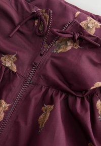 Vêtement bordeaux avec fermeture éclair et cordons, décoré d'un motif répété de têtes de vaches Highland marron clair.
