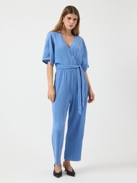 Kvinne som står i en lys blå plissert omslagsjumpsuit med belte i midjen og svarte spisse hæler mot en vanlig hvit bakgrunn.