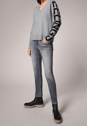 Šedý svetřík s výstřihem do V a černým nápisem na rukávech, v kombinaci se šedými slim-fit džíny a černými kotníkovými botami s masivními podrážkami.