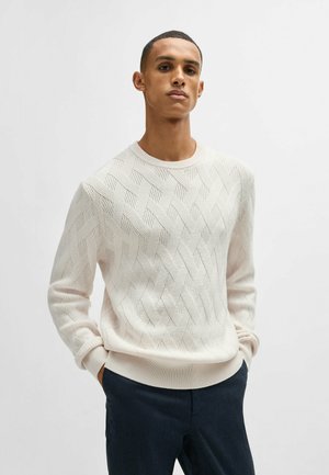 Pullover di cotone chiaro con motivo a diamante testurizzato, scollatura rotonda e orlo a costine. Abbinato a pantaloni scuri.