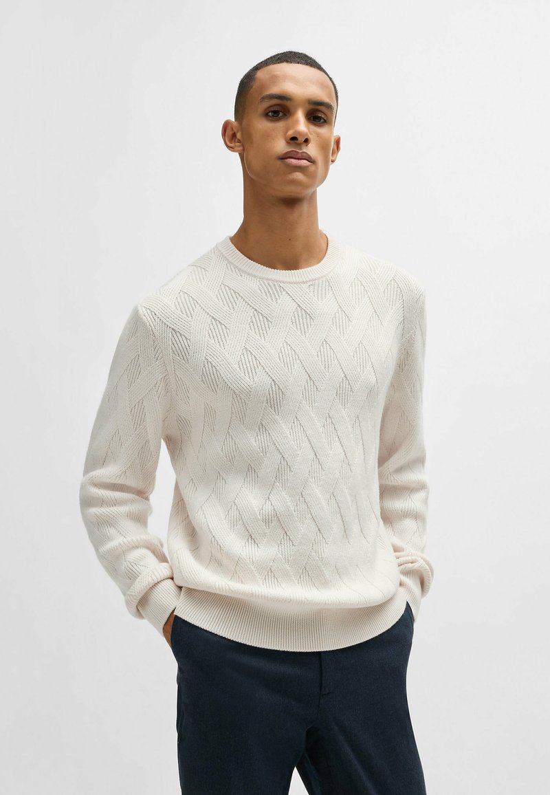 Pullover di cotone chiaro con motivo a diamante testurizzato, scollatura rotonda e orlo a costine. Abbinato a pantaloni scuri.