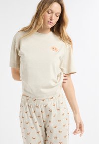 Beige T-Shirt mit kurzen Ärmeln und einem Hundemotiv auf der Brust, kombiniert mit hellen Hosen, die ein wiederholendes Hundemuster und Polka-Dots aufweisen.