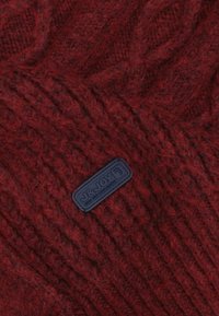 Gebreide rode wollen muts met een textuurpatroon en een blauwe rubberen logo patch met de tekst "J&JOY" aan de zijkant.