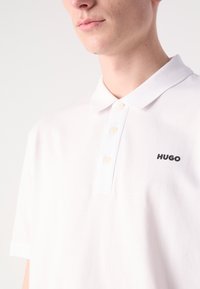 Jeune personne portant un polo blanc à trois boutons avec le logo "HUGO" sur la poitrine gauche.