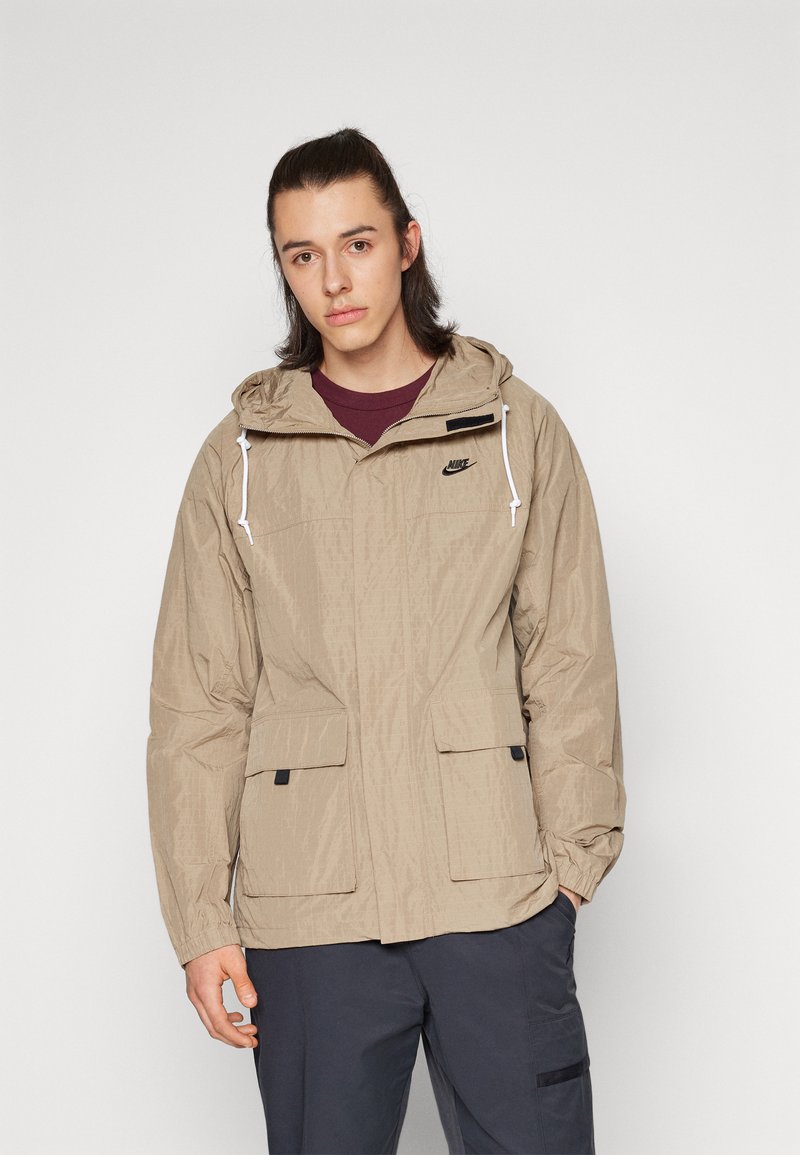 Chaqueta beige de Nike con capucha, que presenta una tela texturizada, dos bolsillos frontales y un logo negro en el pecho. Llevada encima de una camiseta color burdeos.