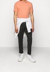 Vaalean oranssi pikeepaita, jossa on kaulus, harmaat istuvat housut ja valkoiset lenkkarit. Neule sidottuna vyötärölle. Suuri tekstuuri, yksinkertainen muotoilu.