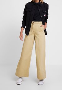 Beżowe spodnie szerokie z kieszeniami z przodu i kontrastowym wykończeniem, w połączeniu z czarną marynarką na guziki i białymi sneakersami.