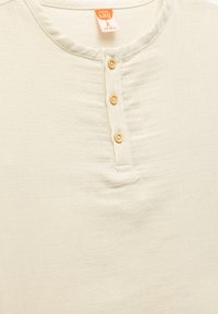 Camisa corta de manga corta en beige claro, hecha de una tela suave, con un escote redondo y detalle de cuatro botones de madera en la parte delantera.